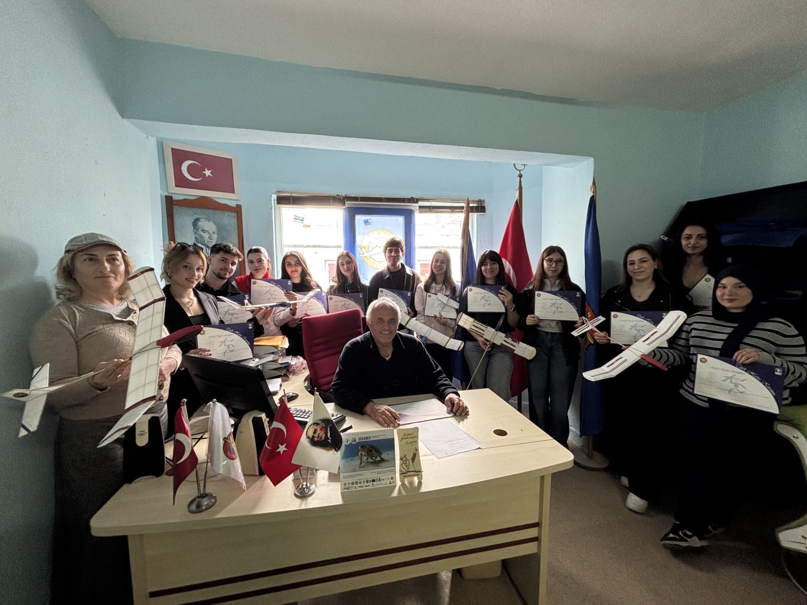 Daha samimi bir ofis ortamında çekilen toplu bir fotoğraf. Cengiz Şatır masasında otururken, etrafında ellerinde hem sertifikalarını hem de yaptıkları model uçakları tutan bir grup öğrenci bulunuyor. Odada Türk bayrağı ve Atatürk portresi yer alıyor.